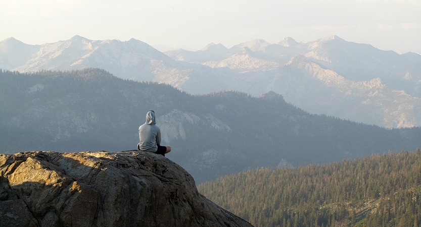 A person faces away from the camera while sitting on a rock, overlooking a mountainous landscape. 